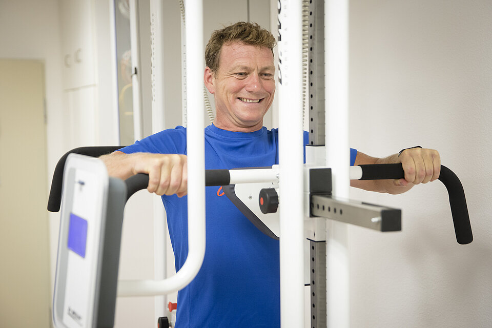 Mann mit blauem T-Shirt in der Sportmedizin sitzt an einem Trainingsgerät und führt eine Kraftübung aus.