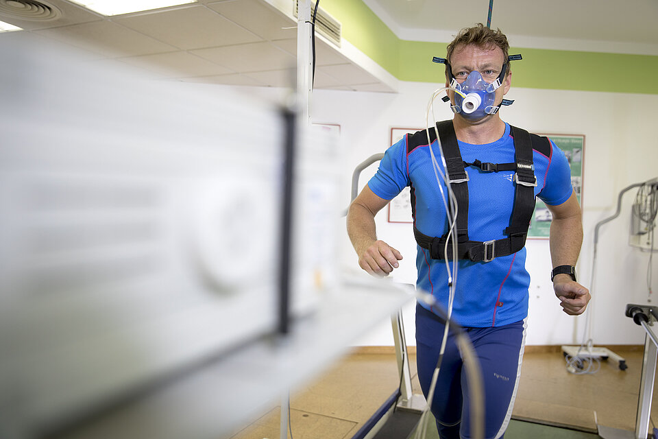 Mann läuft auf dem Laufband während einer Leistungsdiagnostik, verkabelt und mit Sauerstoffmaske, die die Werte misst. Der Hintergrund ist verschwommen, während der Mann im Fokus steht.